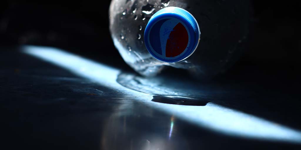 Pepsi Seeking: Tech for Light Weighting of Hot-Fill PET bottles: Image of an empty PET Pepsi bottle on it's side cap facing the camera with the pepsi logo visible and an angular patch of light extending from top left to bottom right of the image